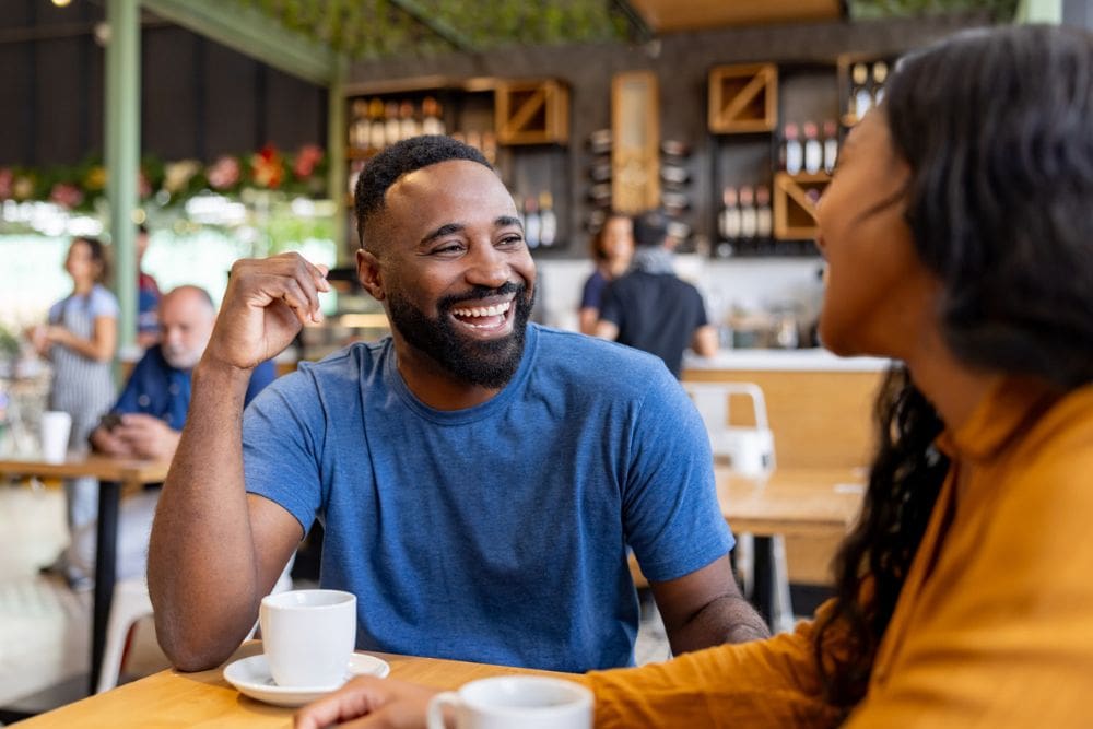 Smiling couple enjoys coffee and conversation at a cozy café, symbolizing healthy social connection and dating in recovery.