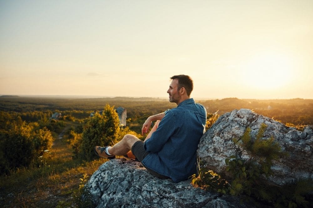 A man sits on a large rock at sunset, looking out over a vast green landscape. He appears relaxed and thoughtful, wearing a denim shirt and shorts, with warm golden light illuminating the scene.