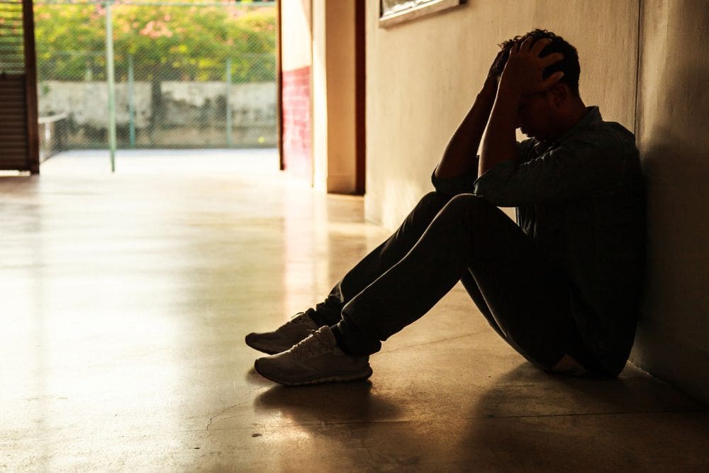 A man sits on the floor in a dim hallway with his head in his hands, appearing overwhelmed or distressed, suggesting emotional struggle or crisis.