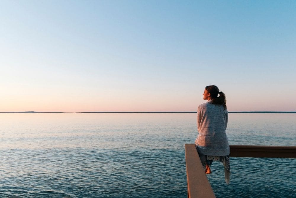 Person sitting on a wooden railing overlooking a calm lake at sunset, wearing a cozy sweater and gazing thoughtfully at the horizon.
