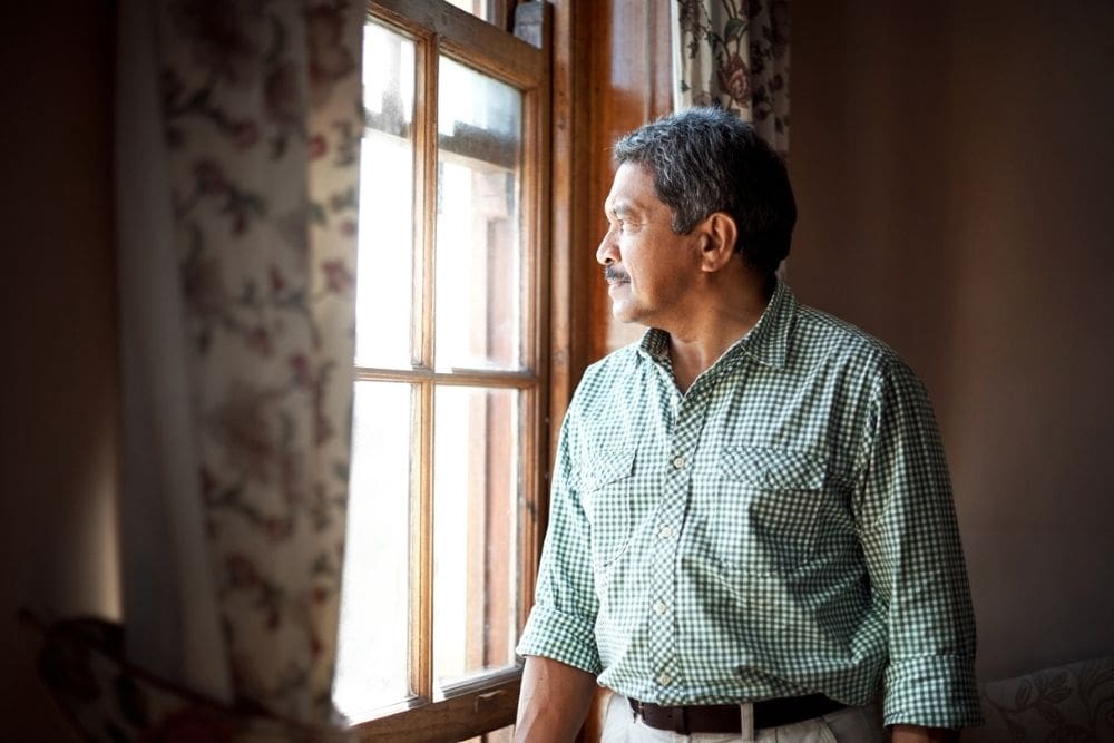 Older man in a checkered shirt standing indoors and gazing thoughtfully out a window