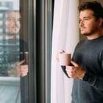 Addiction(1000 x 667 px) Man standing by a window holding a mug, looking outside thoughtfully with his reflection visible in the glass.