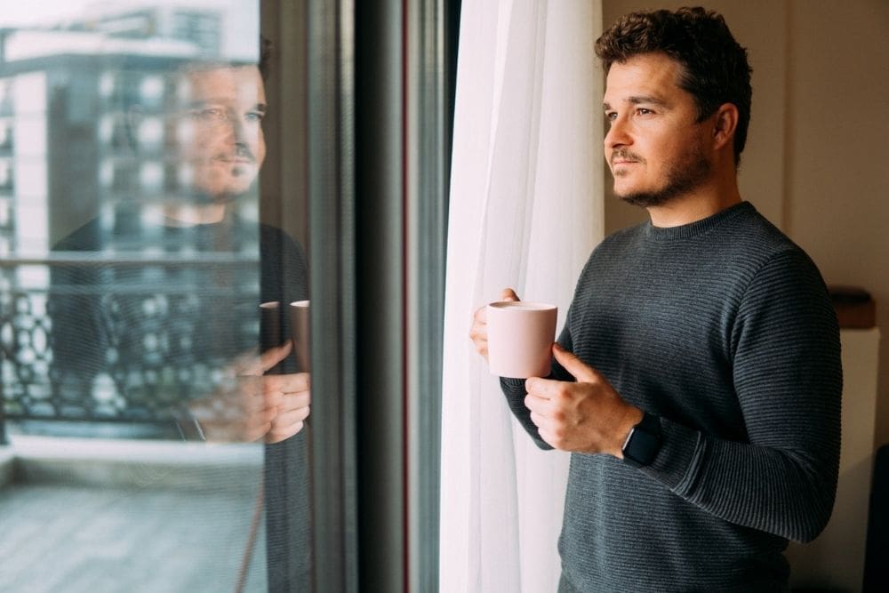 Addiction(1000 x 667 px) Man standing by a window holding a mug, looking outside thoughtfully with his reflection visible in the glass.