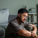 Loneliness in Recovery (1000 x 667 px) A man sits on the floor near a couch with his arms around his knees, looking thoughtful and lonely, reflecting emotional struggles in recovery.