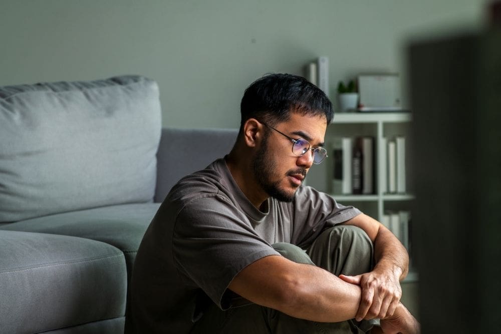 Loneliness in Recovery (1000 x 667 px) A man sits on the floor near a couch with his arms around his knees, looking thoughtful and lonely, reflecting emotional struggles in recovery.