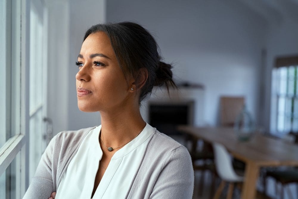 A woman stands indoors looking thoughtfully out of a window, with natural light illuminating her face.