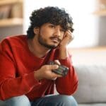 A young man sitting on a couch, holding a TV remote and resting his face on his hand, looking bored or uninterested.