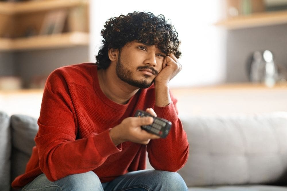 A young man sitting on a couch, holding a TV remote and resting his face on his hand, looking bored or uninterested.