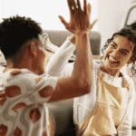 Family Milestones (1000 x 667 px) Two people giving each other a high five while sitting on the floor, celebrating a positive moment at home.
