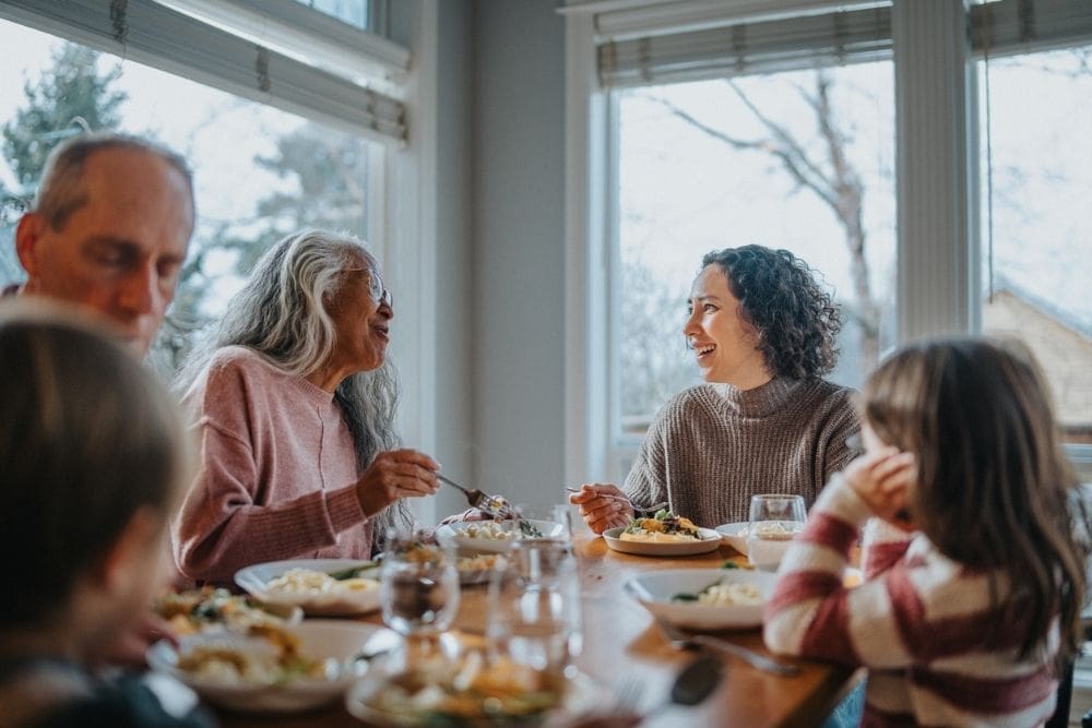 A multigenerational family sits together at a dining table, sharing a meal and conversation in a bright, cozy home setting.