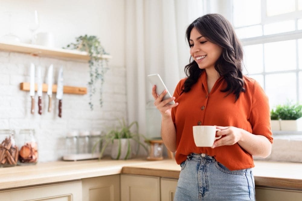Smiling young woman enjoying her coffee while using her smartphone in a homey kitchen setting