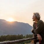 Older adult leaning on a wooden fence, looking out at a mountain landscape during sunset in a quiet, reflective moment.