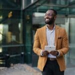 Las Cruces – Tips to Help Rebuild Confidence – Edited Las Cruces - Tips to Help Rebuild Confidence. A yong professional man stands in a modern glass office looking into the distance and smiling, He is wearing a mustard yellow suit with a white button-up shirt holding a white tablet.