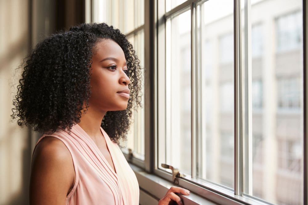 Cravings (1000 x 667) Woman standing indoors, calmly looking out a large window with a thoughtful expression, wearing a sleeveless pink top