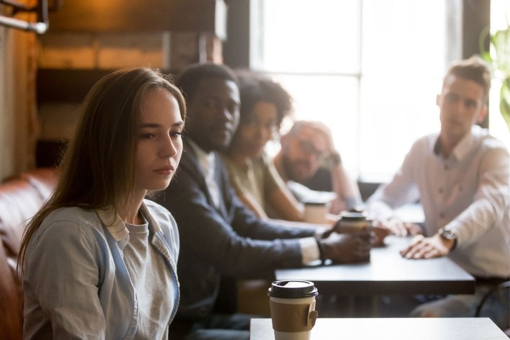 A young woman looks upset and distant while sitting at a table with a group of friends, highlighting feelings of isolation and toxic relationships.