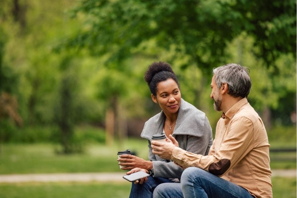 Sponsor Meeting (1000 x 667 px) Two people sitting outdoors on a park bench, holding coffee cups and talking, suggesting a supportive and meaningful conversation.