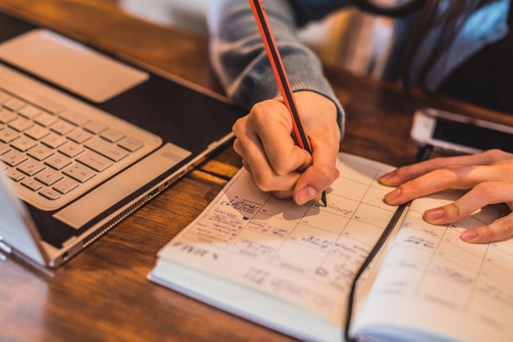 A person writing in a planner next to an open laptop, organizing their schedule and creating a routine.